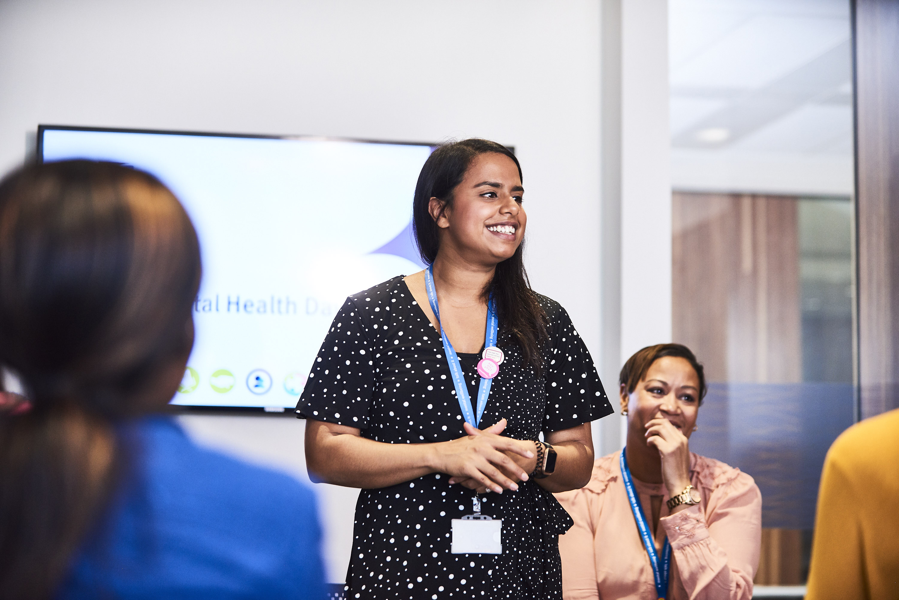Female presenter smiling in front of a group, behind her the screen displays 'World Mental Health Day'